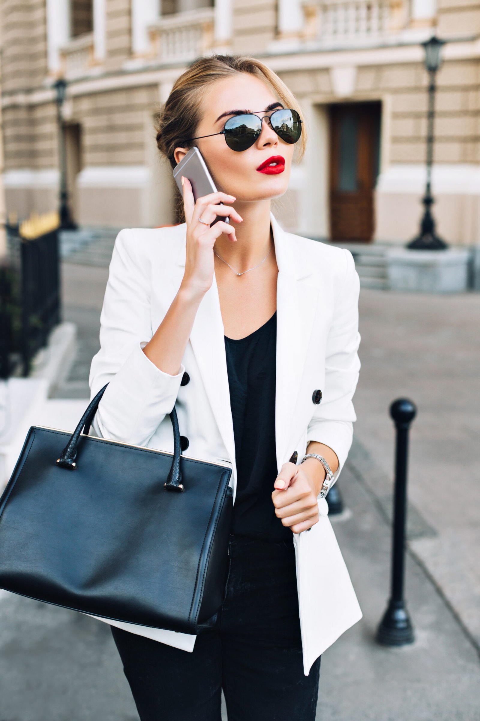 Portrait attractive woman in sunglasses with red lips walking on street . She wears white jacket, black bag, speaking on phone